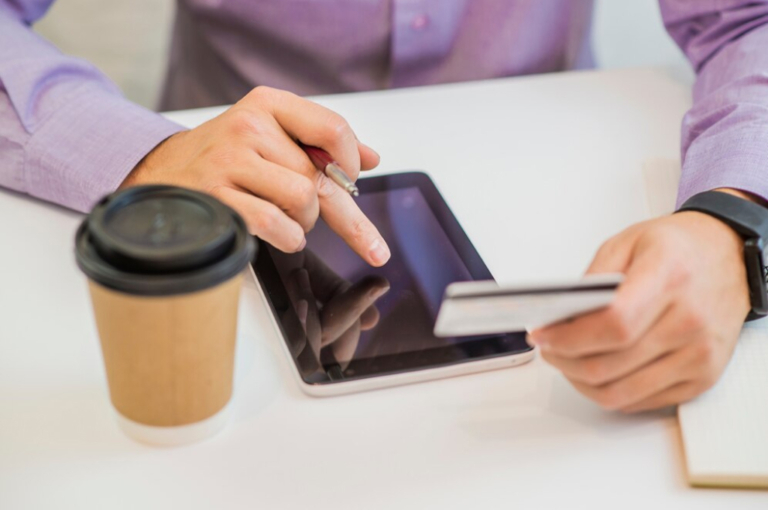 Persona realiza una operación financiera digital con tarjeta y tablet mientras toma café, representando la facilidad del Préstamo por Descuento por Planilla en Coopac San Miguel