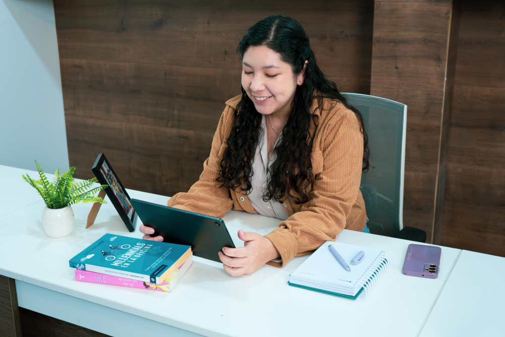 Mujer joven sentada en oficina leyendo desde una tablet, con libros, cuaderno y celular sobre el escritorio, representando el blog de Coopac San Miguel