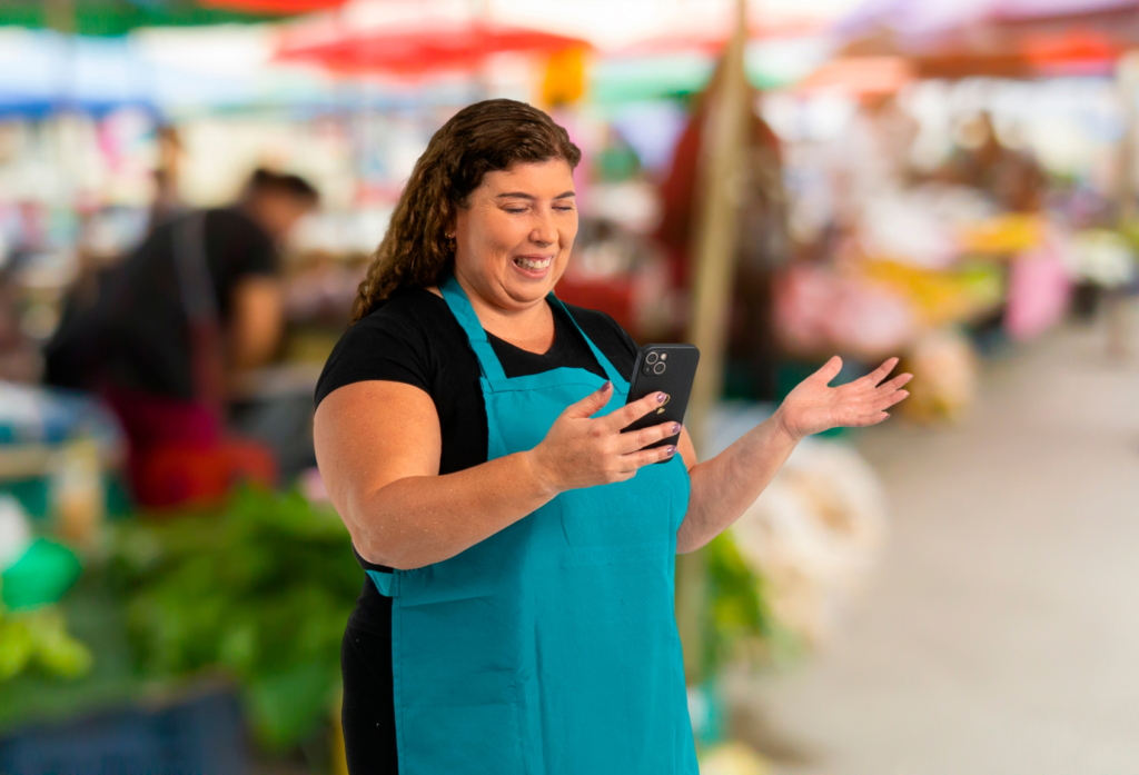 Emprendedora en mercado utilizando su celular para consultar información sobre el Préstamo Emprende Mujer de Coopac San Miguel, en un entorno cotidiano de trabajo