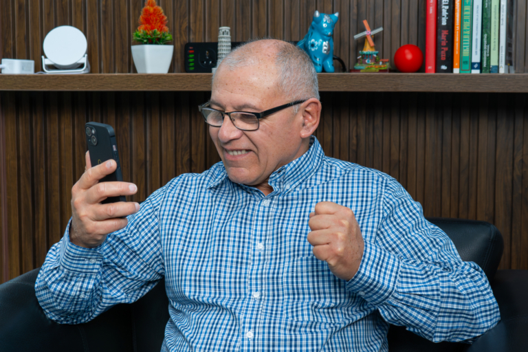Socio mayor sonriente celebrando desde casa al recibir notificación de rendimiento por su Depósito a Plazo Fijo en Coopac San Miguel, reflejando confianza y satisfacción
