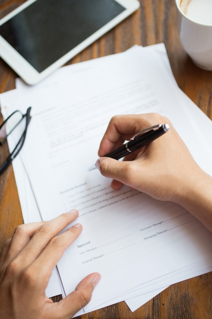 Persona llenando formulario de solicitud sobre escritorio con documentos, tablet y taza de café