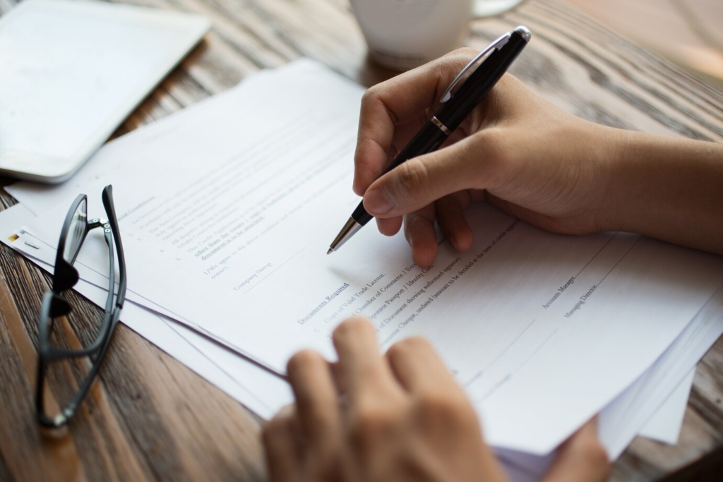 Manos de un hombre de negocios firmando documentos sobre una mesa de madera con lentes, café y una tablet al costado en un ambiente de oficina